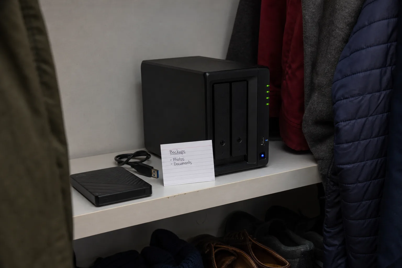 A two-bay network attached storage enclosure on a shelf in a hall closet, with a bare external hard drive and a USB cable beside it, and a handwritten index...