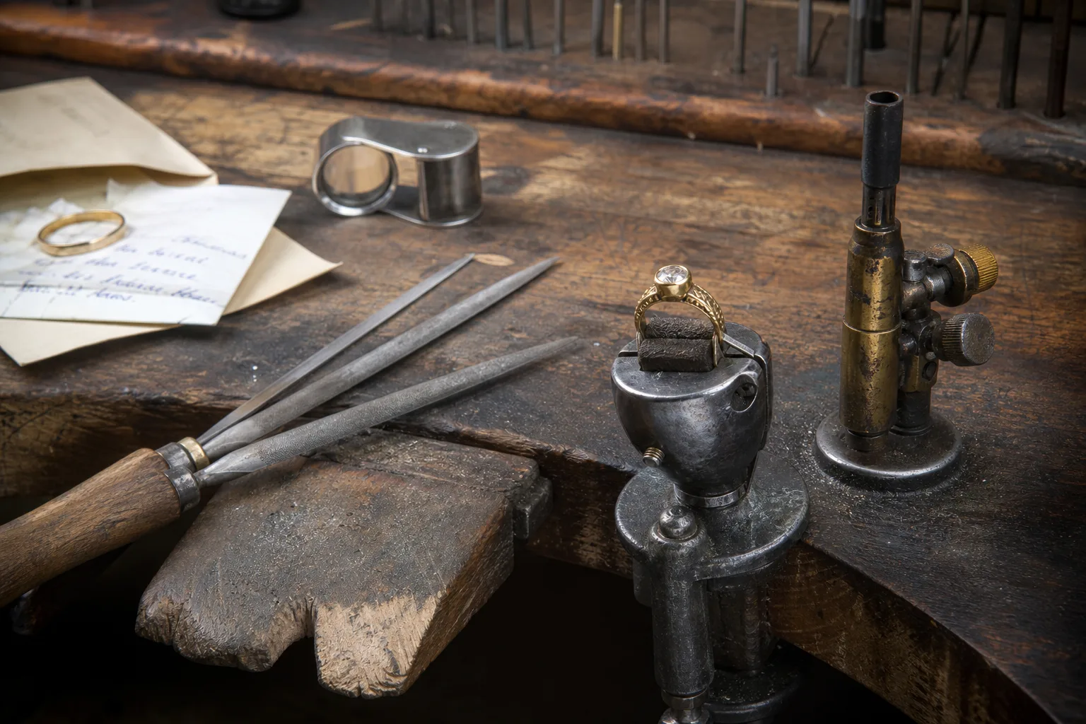 A jeweler's bench with a gold ring held in a ring clamp beside a small torch, files, a loupe and a handwritten repair envelope
